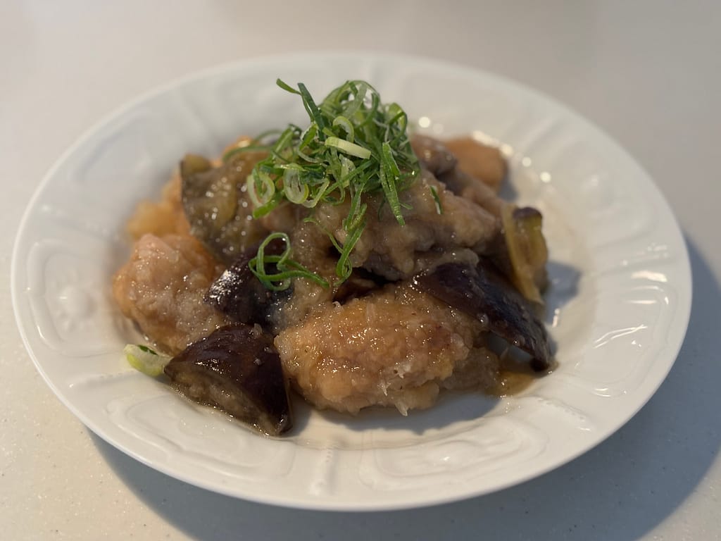 とろみあんが絡んだ鶏肉とナスの煮物。白い皿に盛りつけられ、青ねぎが彩りを添えている。