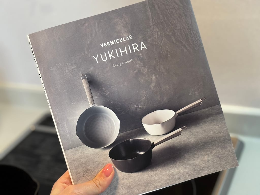 バーミキュラ ユキヒラの公式レシピブックを手に持った写真。スタイリッシュな鍋のビジュアルが表紙にデザインされている