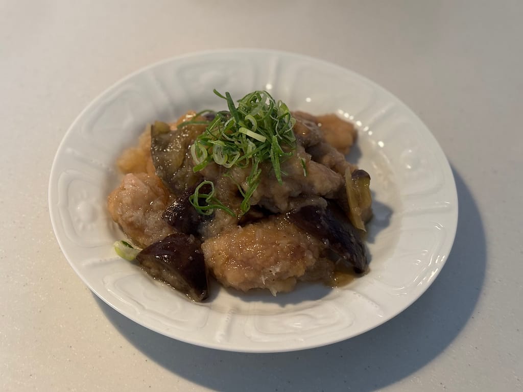 とろみあんが絡んだ鶏肉とナスの煮物。白い皿に盛りつけられ、青ねぎが彩りを添えている。