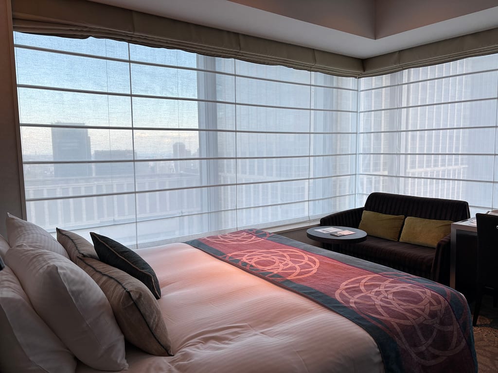コーナールームのベッドと窓「ホテルメトロポリタン丸の内のコーナールーム。大きな窓から東京の街並みを一望でき、ベッドとソファが配置されている。」