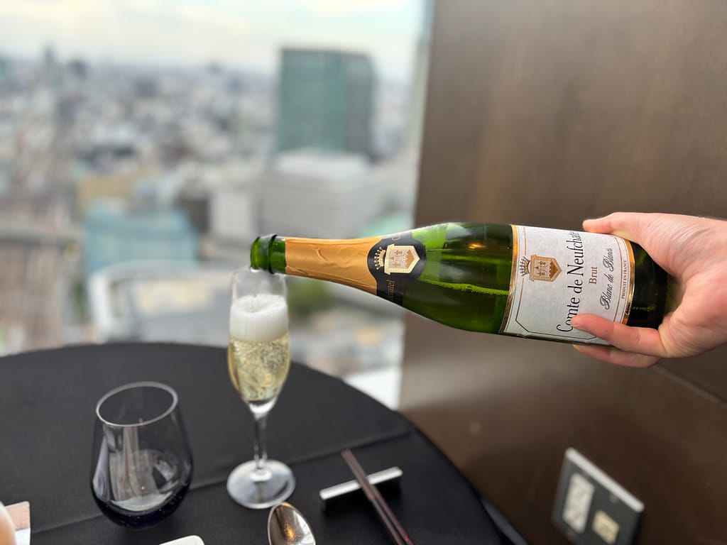 スパークリングワインを注ぐ様「Comte de Neuchatelのスパークリングワインをグラスに注ぐ手元。背景には高層ビル群が広がる。」