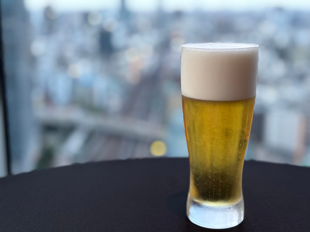 ビール「東京の街並みを背景に、きめ細かな泡がのった生ビールのグラス。」