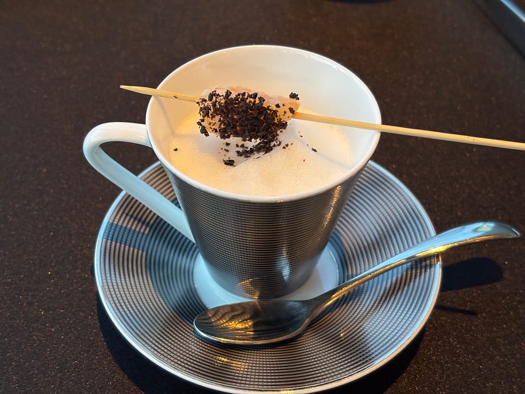 スープ仕立ての一品「カップに注がれた泡立ちスープ。上には串に刺した具材と黒いトッピングが飾られている。」