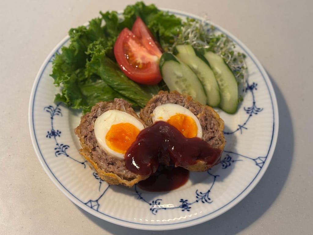 盛り付けた完成料理（アップ）
「半熟卵が入ったスコッチエッグを半分に切り、トマトソースをかけて野菜サラダと一緒に盛り付けた一皿」
