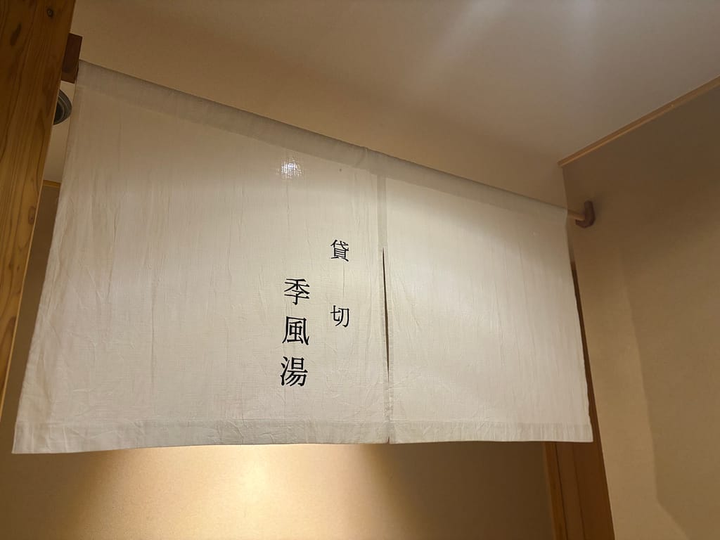 貸切風呂「季風湯」暖簾。吉田屋の貸切風呂「季風湯」の暖簾。白い布にシンプルな文字で温泉の入り口を示す落ち着いた雰囲気。